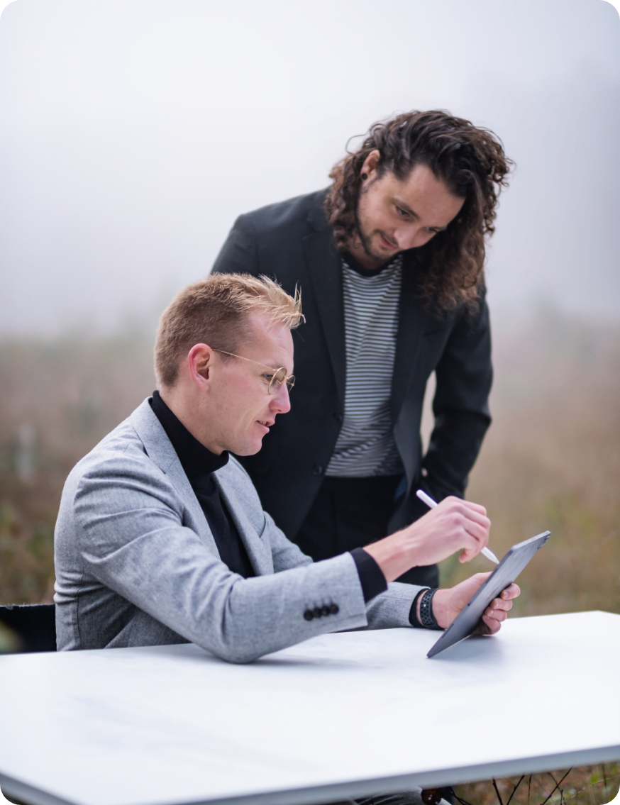 Twee mannen, de één zittend en de ander staand, zijn gefocust op een digitale tablet. Ze zijn buiten, mogelijk in een mistig veld, gekleed in zakelijke, casual kleding. De staande man heeft krullend haar