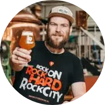 Een bebaarde man met een pet en een zwart t-shirt met de tekst "rock on hard rock city" houdt een glas amberkleurig bier omhoog, staande in een brouwerij met brouwapparatuur op de achtergrond.